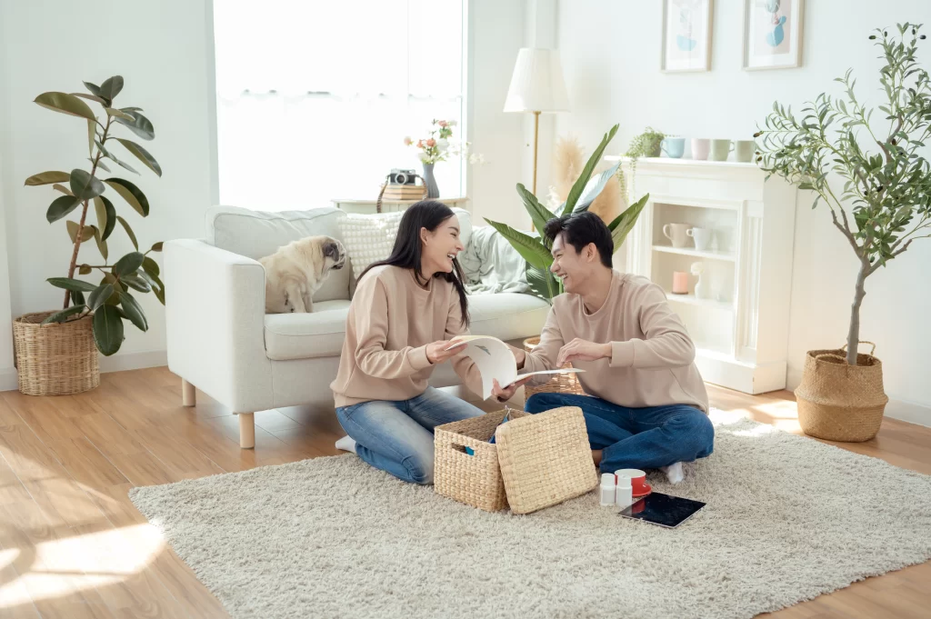 Couple Smiling While Carrying Plants and Baskets in a Sunlit Living Room Filled With Greenery seguro-de-hogar-inicio